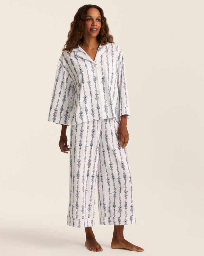 A woman stands barefoot against a plain light background wearing the Greek Embroidered Olivia Wide Leg Pima Pajama Set in Navy Mayfair Toile—a loose-fitting white pajama set adorned with blue vine-like vertical stripes.
