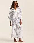 A woman stands barefoot against a plain light background wearing the Greek Embroidered Olivia Wide Leg Pima Pajama Set in Navy Mayfair Toile—a loose-fitting white pajama set adorned with blue vine-like vertical stripes.