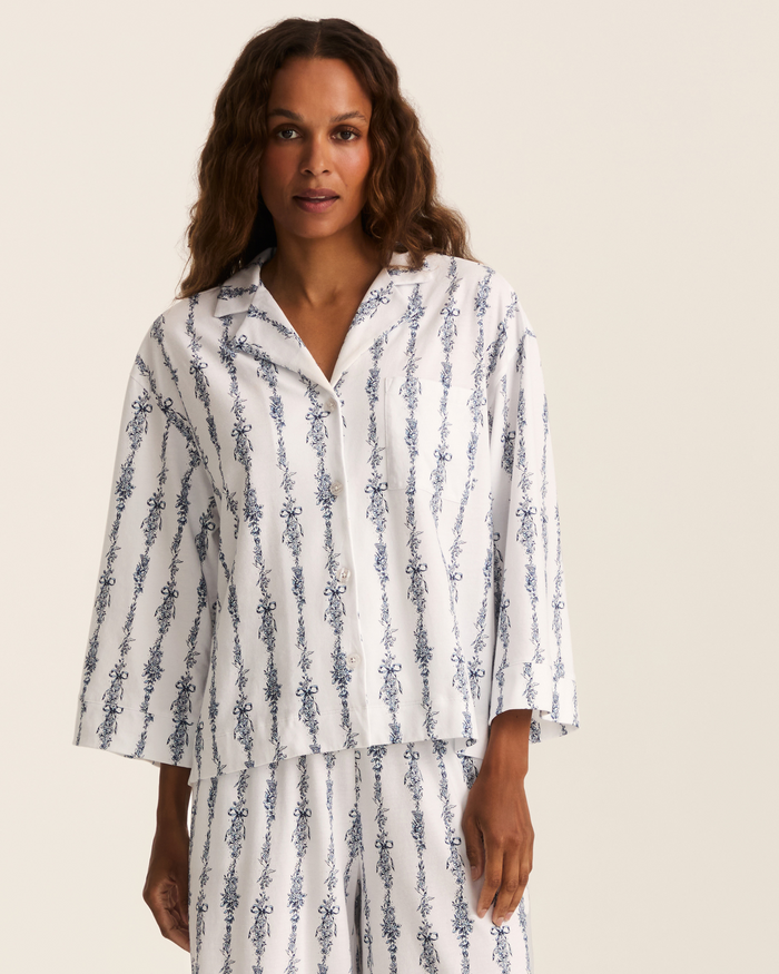 A woman stands against a plain background, looking relaxed at the camera, wearing the Greek Embroidered Olivia Wide Leg Pima Pajama Set in Navy Mayfair Toile.