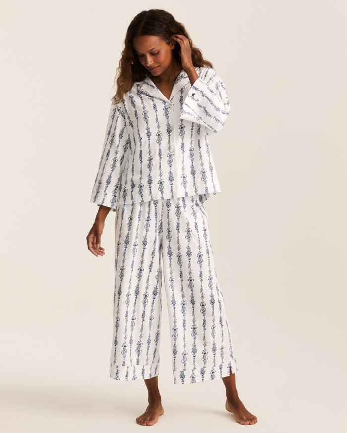 A woman stands barefoot against a plain background, wearing the Greek Embroidered Olivia Wide Leg Pima Pajama Set in Navy Mayfair Toile, which features blue vertical patterns on a matching white set with long sleeves and wide-leg pants.
