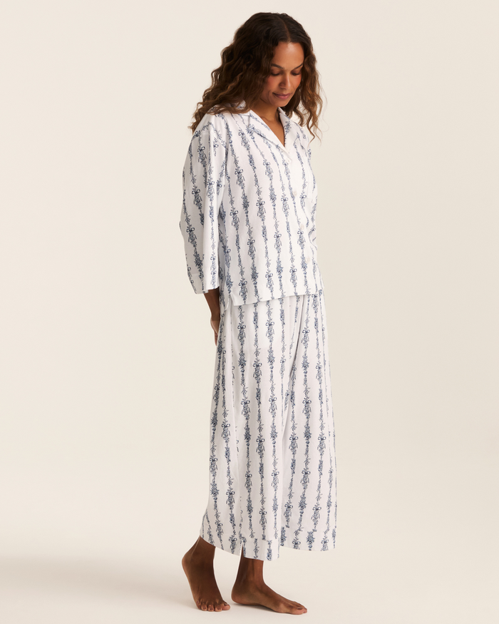A woman stands barefoot against a simple light background, wearing the Greek Embroidered Olivia Wide Leg Pima Pajama Set in Navy Mayfair Toile. The matching set features a long-sleeved button-up top and wide-leg pants, both adorned with elegant navy floral stripes. She gazes down gently, showcasing the pajamas’ refined style.