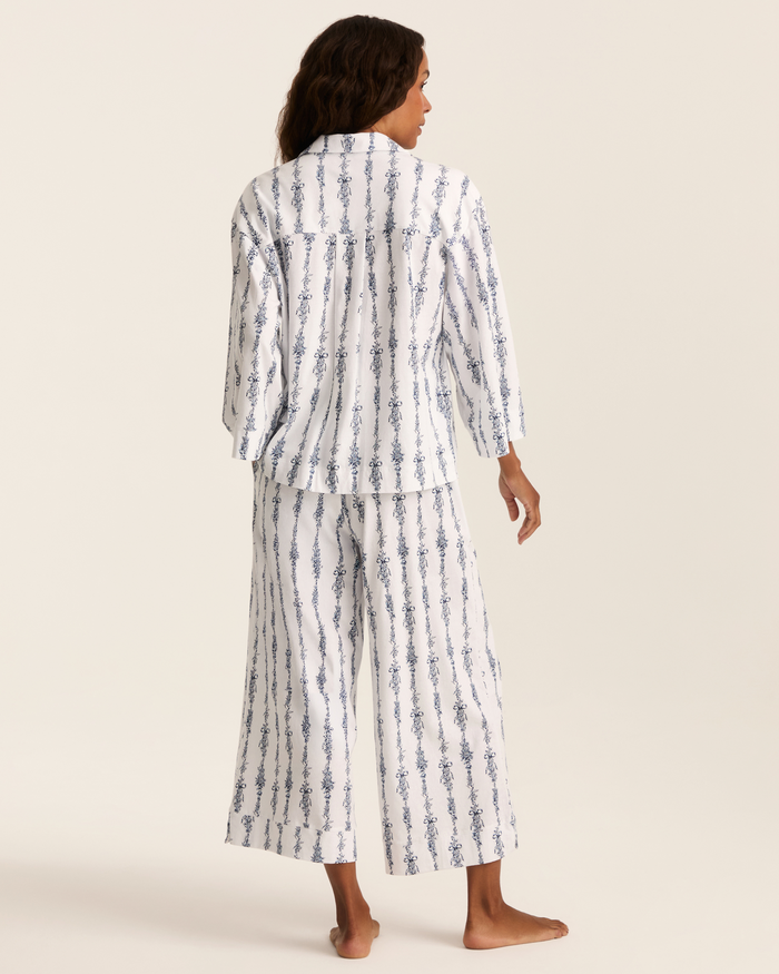 A person stands barefoot facing away, wearing the Greek Embroidered Olivia Wide Leg Pima Pajama Set in Navy Mayfair Toile, which features matching white pajama pants and a top adorned with vertical blue floral stripes. The background is plain and light-colored.
