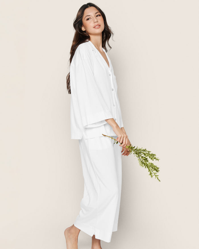 Wearing the Greek Embroidered Olivia Wide Leg Pima Pajama Set in White, a woman with long dark hair stands barefoot against a beige background, gently holding a small bouquet of green foliage and displaying a relaxed, serene expression.