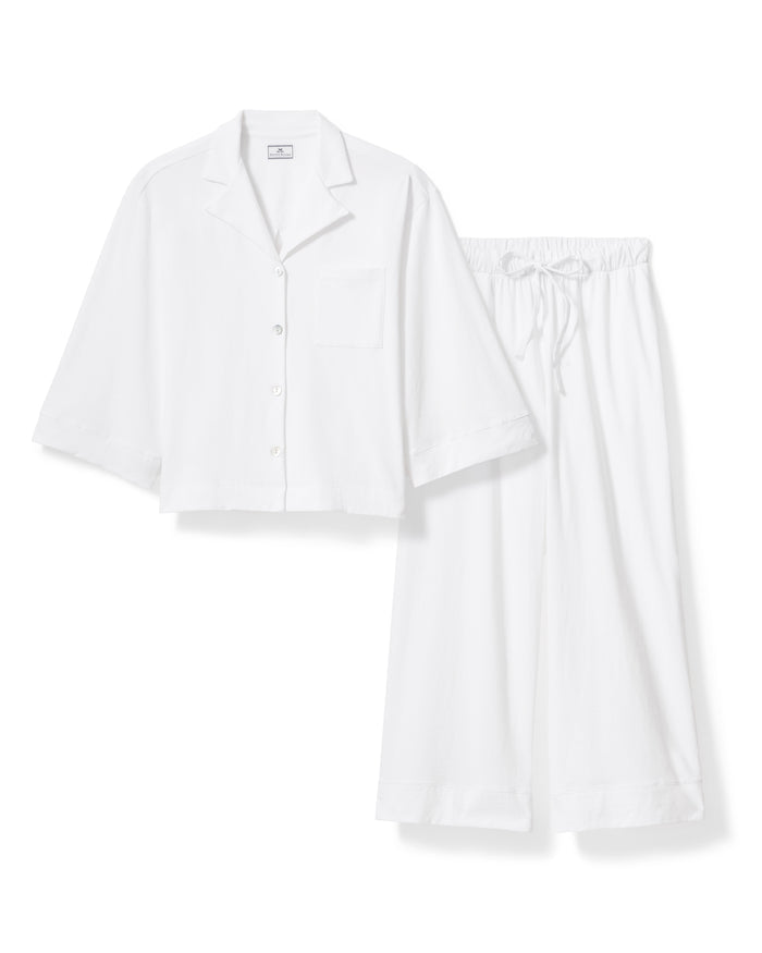 A person wears The Olivia Wide Leg Pima Pajama Set in White by Petite Plume, showcasing a luxurious Peruvian Pima cotton ensemble with a loose-fitting long-sleeve buttoned shirt and wide-leg pants. They hold a branch with green leaves, standing barefoot against a light background.
