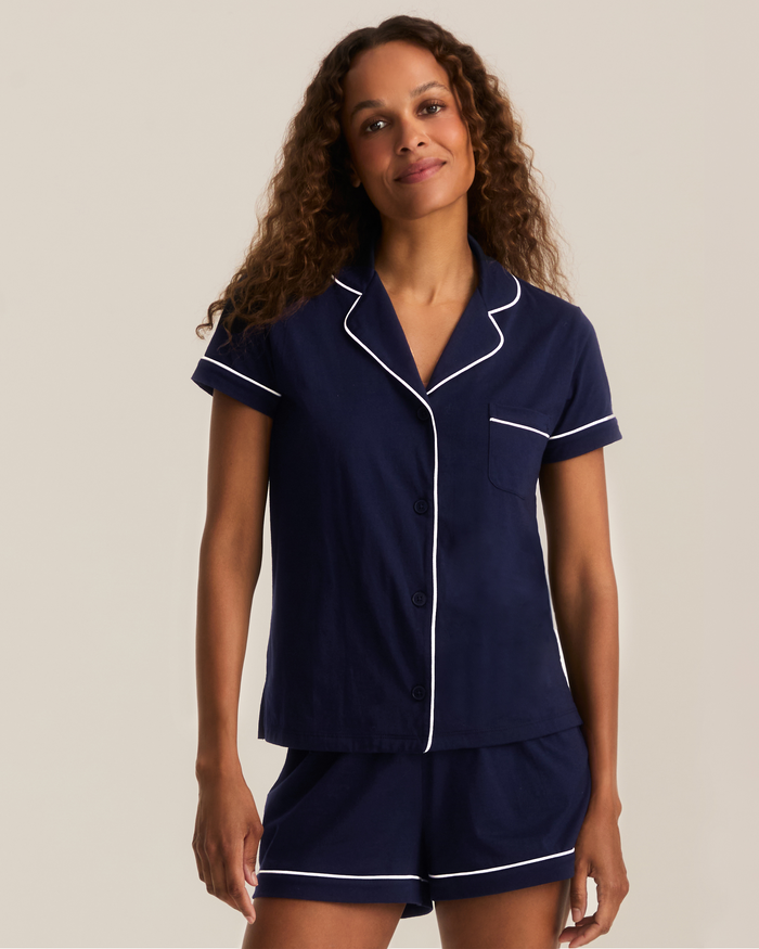 A woman wearing the Greek Embroidered Poppy Lightweight Pima Short Set in Navy, featuring white piping, stands and smiles against a plain, light background.