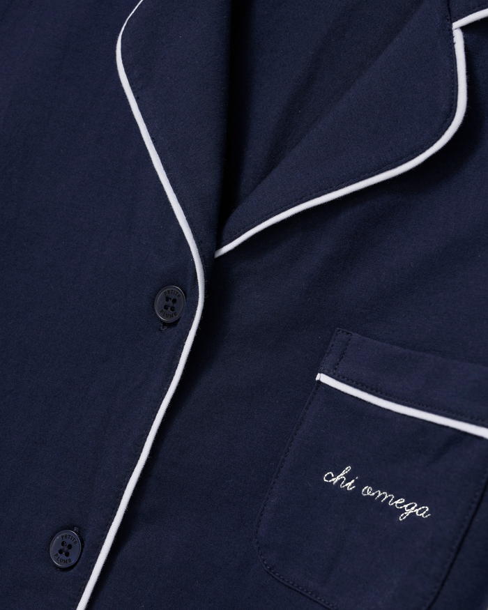 Close-up of the Greek Embroidered Poppy Lightweight Pima Short Set in Navy, featuring a navy blue pajama top with white piping. The pocket is embroidered with “chi omega” in white cursive, and two black buttons are visible.