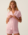 A woman wearing The Poppy Lightweight Pima Short Set in Pink, featuring a light pink color with white trim and short sleeves, stands against a plain light background, smiling slightly while holding the collar of her top.