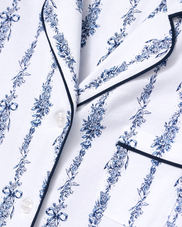 Close-up of the Greek Embroidered Poppy Pima Short Set in Navy Mayfair Toile, featuring a white pajama top with blue floral and bow patterns, white buttons, black piping, and a chest pocket.