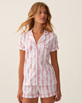 A woman wears The Poppy Pima Short Set in Pink Mayfair Toile, featuring a matching short-sleeve pajama set with a pink and white floral stripe pattern, standing against a plain light background.