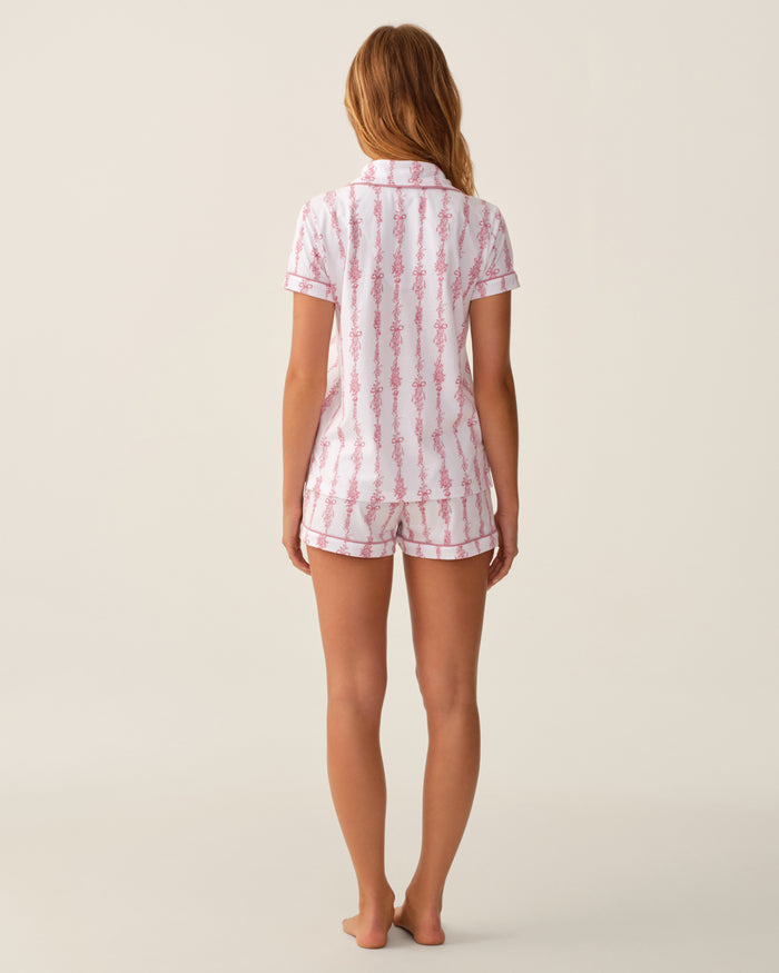 A woman stands barefoot facing away, wearing The Poppy Pima Short Set in Pink Mayfair Toile—a matching short-sleeve button-up top and shorts pajama set with a pink and white pattern—against a plain light background.