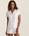 A woman with wavy hair wears The Poppy Pima Short Set in White with Pink Piping, featuring a short-sleeved pajama top with front buttons and matching shorts. She stands facing forward and smiles softly.
