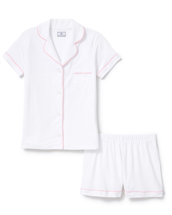 The Poppy Pima Short Set in White with Pink Piping is displayed neatly on a plain white background, featuring a short-sleeved, button-down pajama top with pink piping and a chest pocket, paired with matching white shorts accented by pink trim and an elastic waistband.