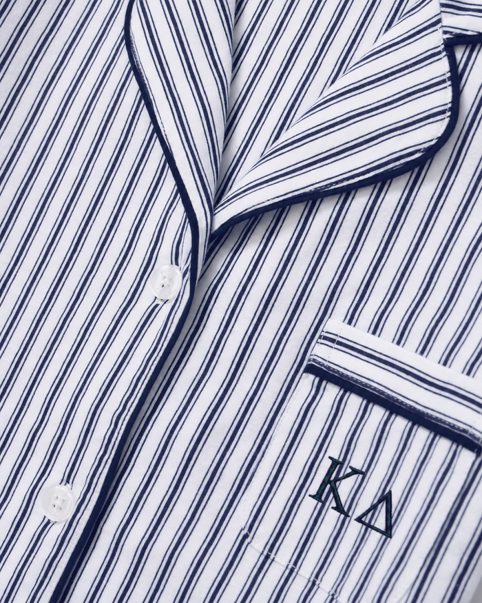 Close-up of the Greek Embroidered Poppy Lightweight Pima Short Set in Navy French Ticking, featuring a white pajama shirt with thin navy stripes, white buttons, navy piping, and a pocket embroidered with KΔ. The smooth fabric appears neatly pressed.