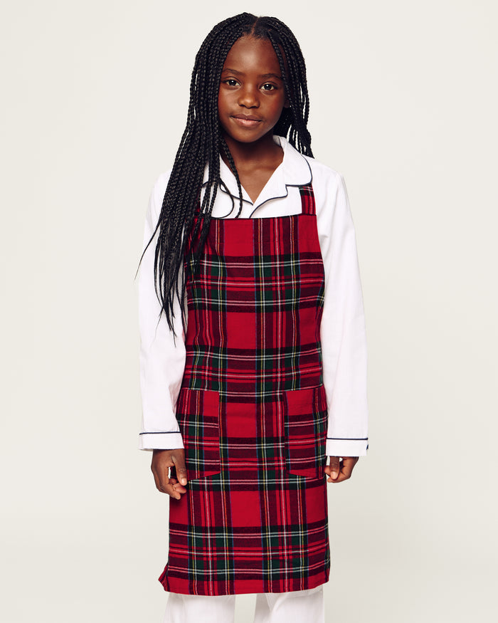 Two Petite Plume Kids Aprons in Imperial Tartan with pockets hang on a wooden peg rack, alongside a white dish towel with red and black patterns. A wooden spoon and spatula peek from one apron pocket, perfect for holiday baking against a light brown backdrop.