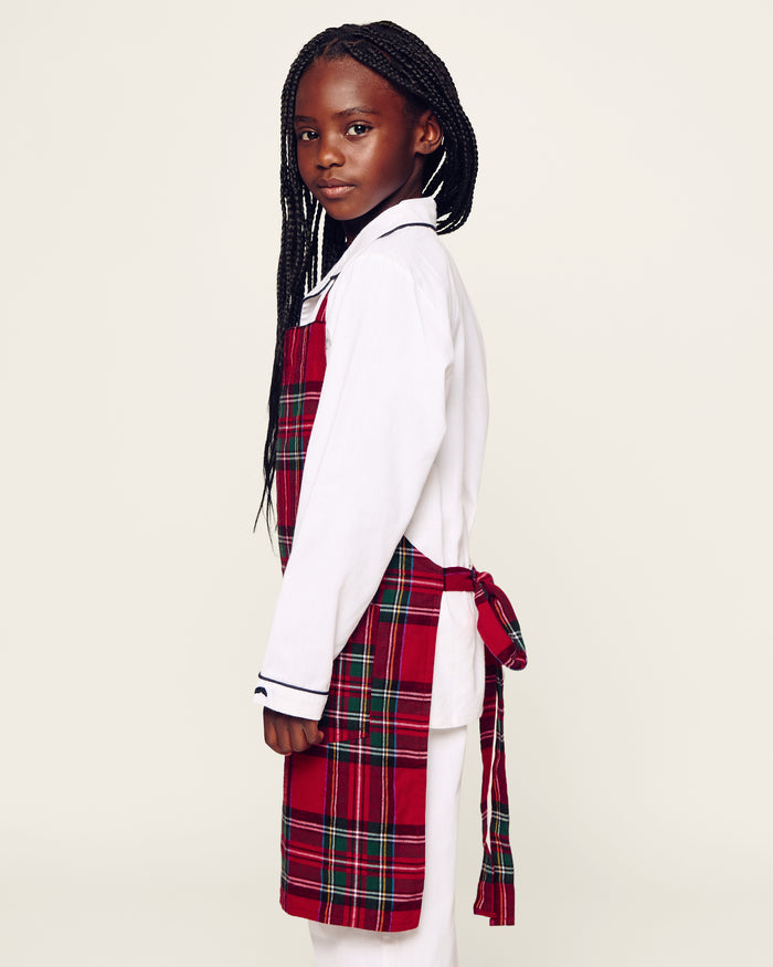 Two Petite Plume Kids Aprons in Imperial Tartan with pockets hang on a wooden peg rack, alongside a white dish towel with red and black patterns. A wooden spoon and spatula peek from one apron pocket, perfect for holiday baking against a light brown backdrop.