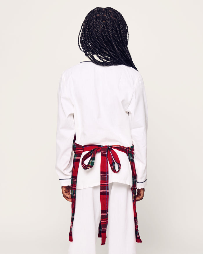 Two Petite Plume Kids Aprons in Imperial Tartan with pockets hang on a wooden peg rack, alongside a white dish towel with red and black patterns. A wooden spoon and spatula peek from one apron pocket, perfect for holiday baking against a light brown backdrop.