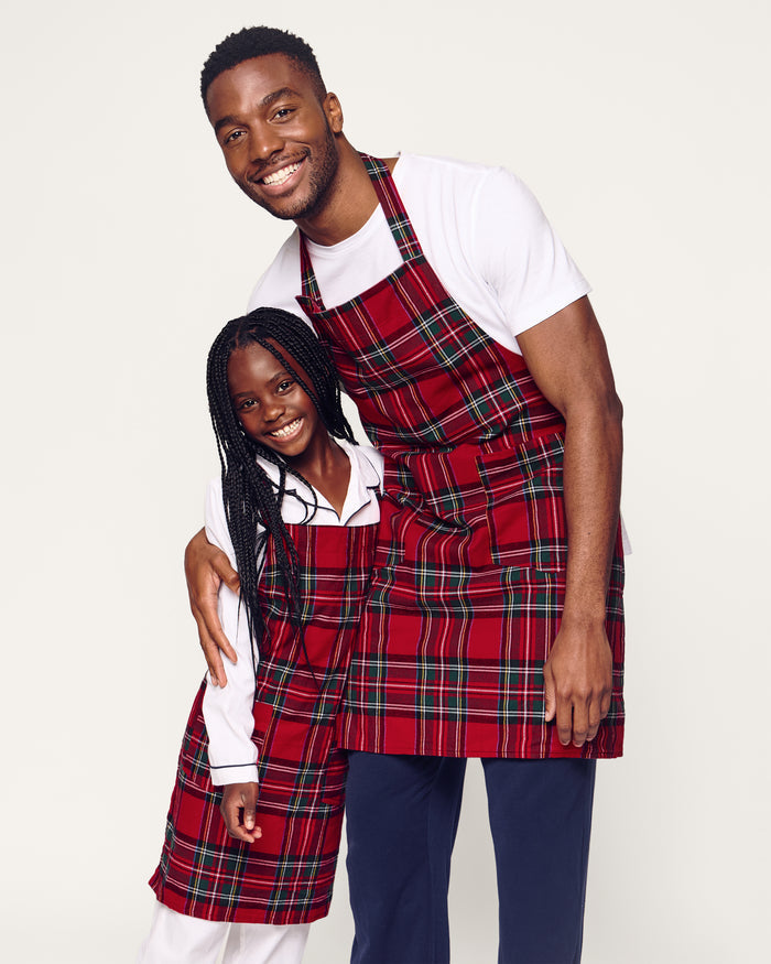 Two Petite Plume Kids Aprons in Imperial Tartan with pockets hang on a wooden peg rack, alongside a white dish towel with red and black patterns. A wooden spoon and spatula peek from one apron pocket, perfect for holiday baking against a light brown backdrop.