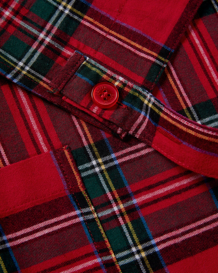 Two Petite Plume Kids Aprons in Imperial Tartan with pockets hang on a wooden peg rack, alongside a white dish towel with red and black patterns. A wooden spoon and spatula peek from one apron pocket, perfect for holiday baking against a light brown backdrop.