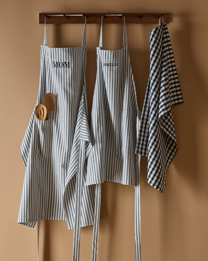 Two navy blue and white striped Petite Plume aprons hang on a wooden rack against a beige wall, ideal for holiday baking. The left one, labeled Mom, holds two wooden spoons. In the middle, a Daniela Kids Apron in Navy French Ticking hangs next to a matching striped dish towel.