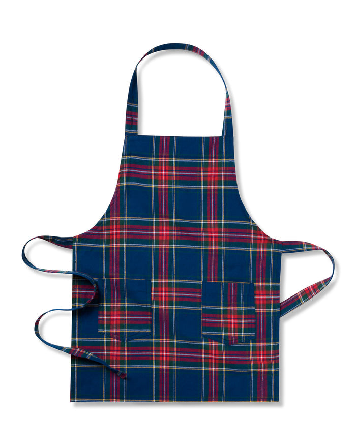 Two Windsor tartan kids aprons from Petite Plume hang on a wooden rack next to a striped dish towel, evoking holiday baking nostalgia. One apron, embroidered with Dad, holds a wooden spoon in its pocket against the warm beige wall backdrop.