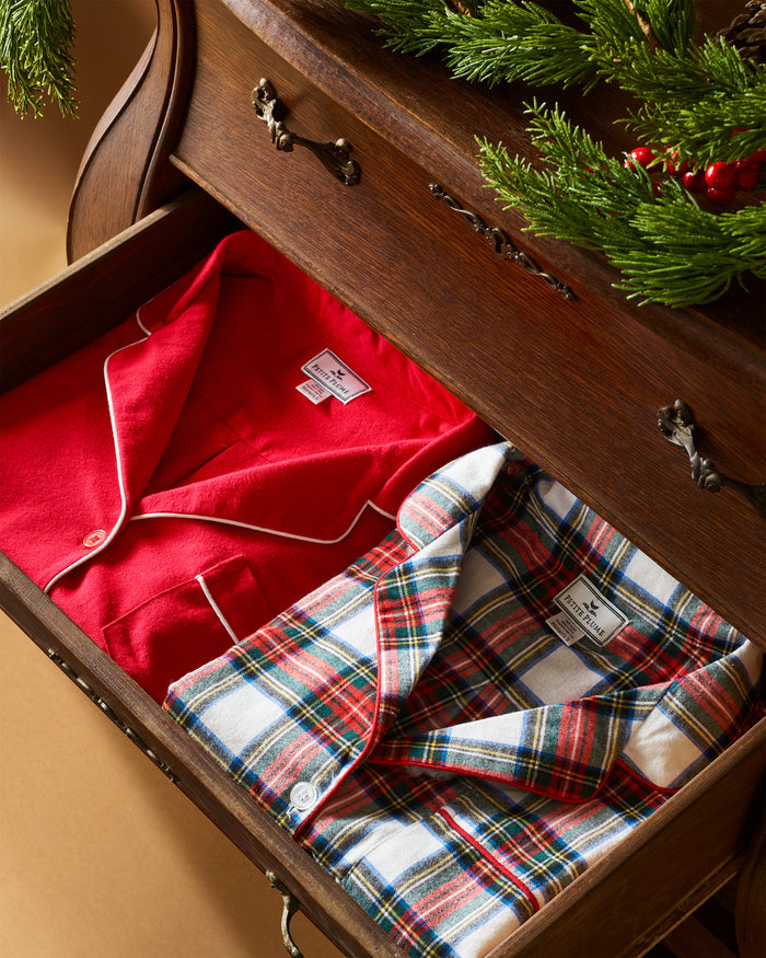 A wooden drawer is partially open, showing two neatly folded pajamas: a Petite Plume classic twill set in red with white piping and a Kids Brushed Cotton Set in Balmoral Tartan. A festive evergreen garland with red berries decorates the dresser top.