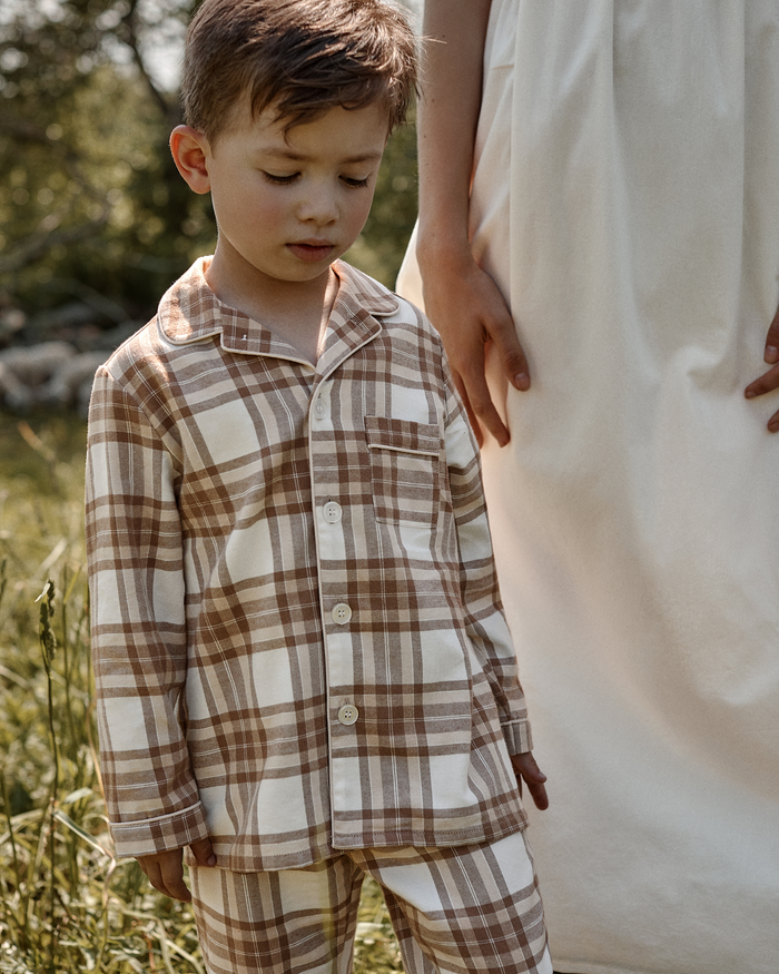 A young boy wears the Kid’s Twill Pajama Set in Cotswolds Tartan as he stands outdoors beside an adult in a white dress, looking down amid lush greenery.
