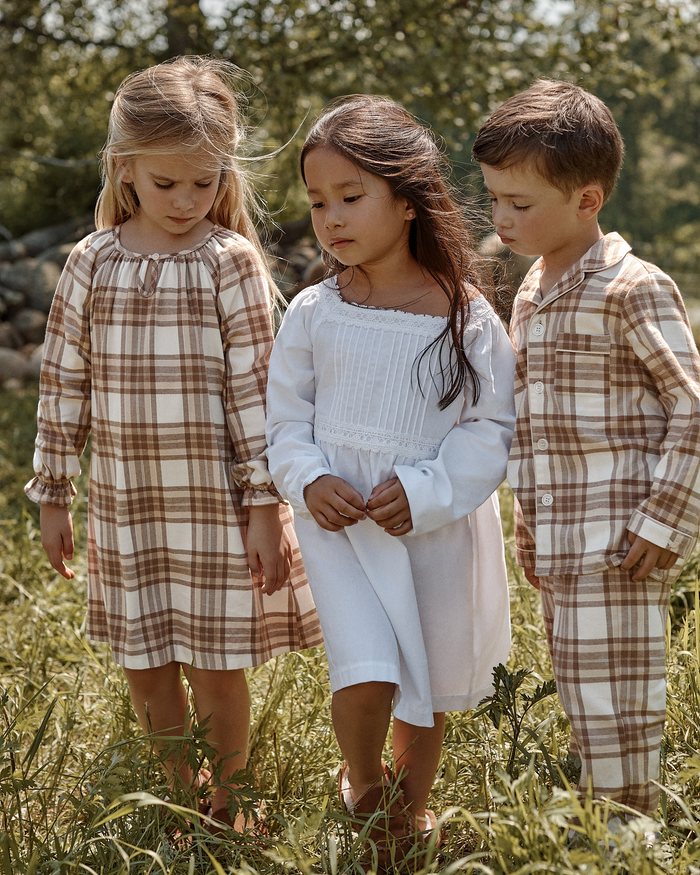 Three children stand together in a grassy outdoor setting. Two wear the Kid’s Twill Pajama Set in Cotswolds Tartan, featuring beige plaid, while one wears a white dress. Sunlight filters through the trees in the background.