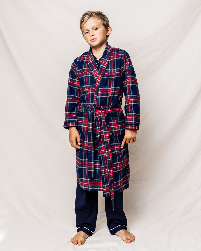 A young boy stands barefoot, wearing the Kid’s Brushed Twill Robe in Windsor Tartan over dark pajamas, looking at the camera with a neutral expression against a plain white backdrop.