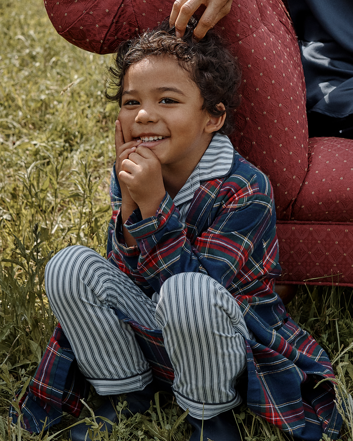 A smiling child in striped pajamas and the Kid’s Brushed Twill Robe in Windsor Tartan crouches on grass beside a red patterned chair, while an adult’s hand rests gently on the child’s head.