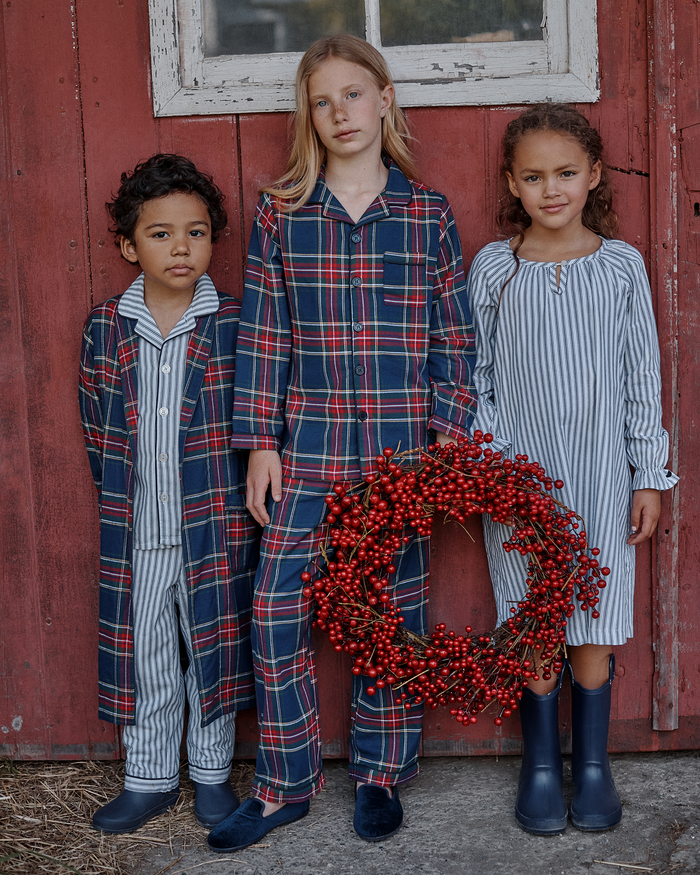 Three children wearing Kid’s Brushed Twill Robes in Windsor Tartan and rain boots stand in front of a red wooden wall. The child in the center holds a large wreath made of red berries. All look toward the camera with neutral expressions.