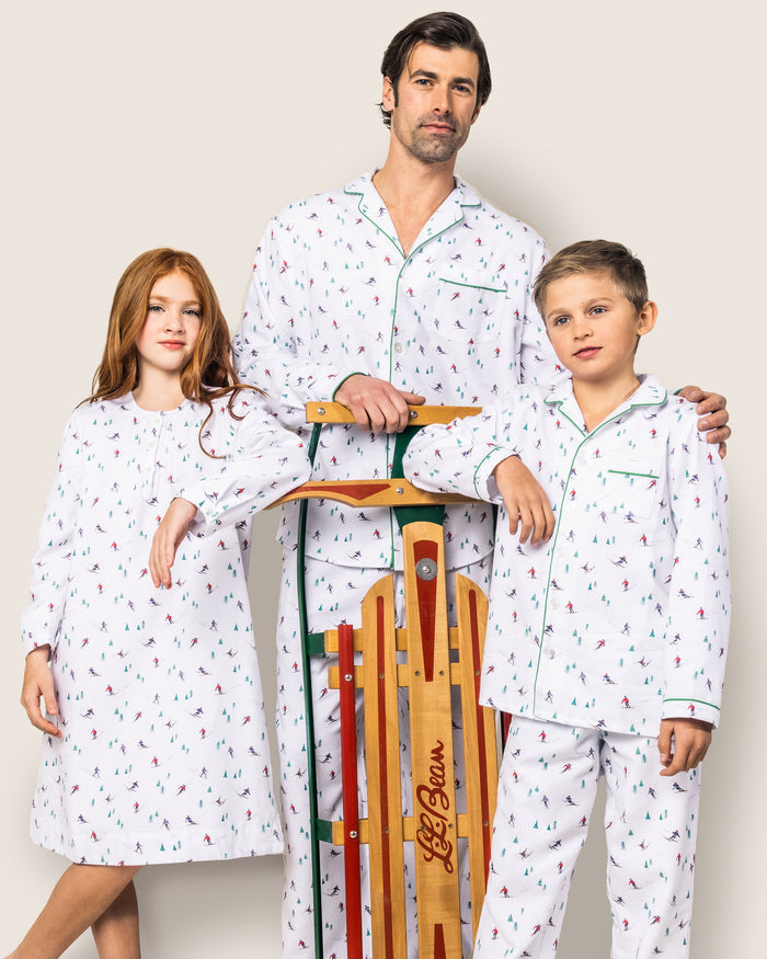 A man, a young girl, and a boy wear matching Petite Plumes Kids Flannel Pajama Set in Après Ski print, safe with flame-retardant fabric. The man and boy rest their hands on a wooden sled against a neutral background.