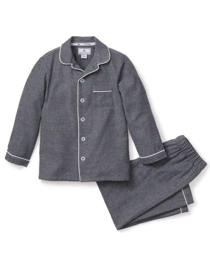 The Kids Flannel Pajama Set in Grey by Petite Plume offers a cozy design with white piping, featuring a button-up top with a chest pocket and pants with an elastic waistband. Made from soft, flame-retardant fabric, it ensures both comfort and safety.