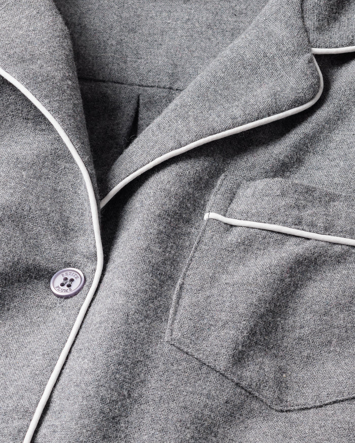 Close-up of Petite Plumes Kids Flannel Pajama Set in Grey, featuring textured fabric, white piping, a front pocket, and a button near the lapel. It is crafted from flame-retardant material for safety and resembles a childrens sleepwear design.