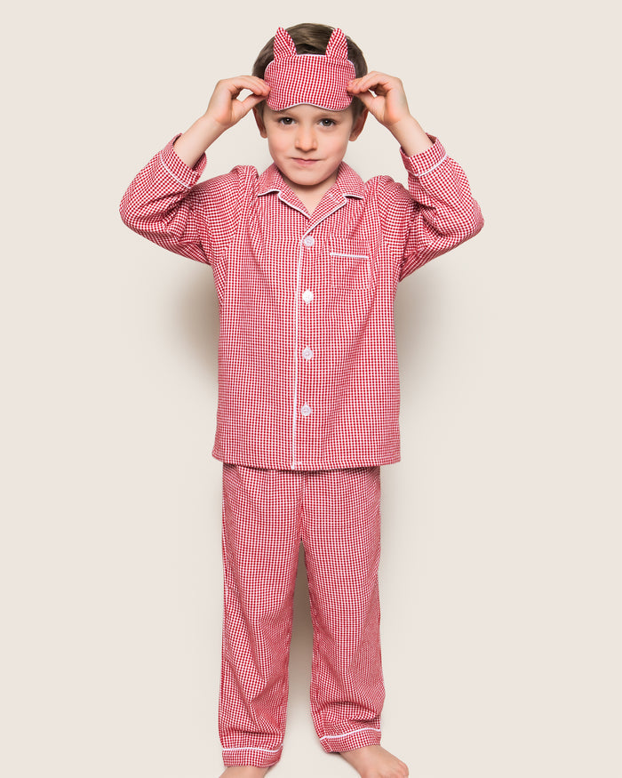 A child, in Petite Plumes Red Mini Gingham Kids Flannel Pajama Set, stands barefoot against a neutral background adjusting a matching sleep mask.