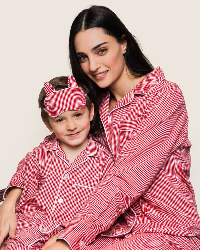 A woman and child happily sit together in Petite Plumes matching flannel pajamas, with the child sporting a Red Mini Gingham sleep mask. They embody the warmth of a classic set against a plain backdrop.
