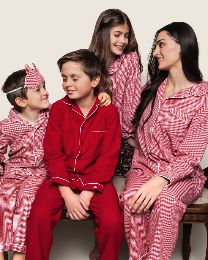 A family of four in matching Petite Plume pajamas sits on a bench. The mother and two kids wear the Kids Flannel Pajama Set in Red Mini Gingham, while the boy in the center dons a solid red set. They smile warmly at each other, radiating togetherness.