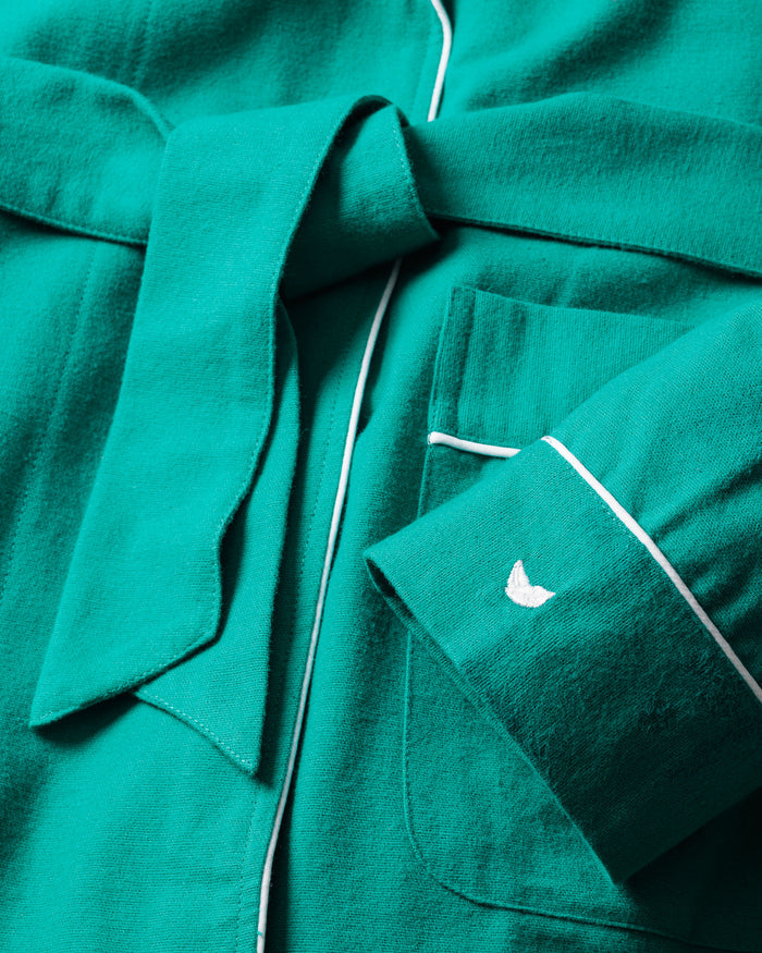 The Petite Plume Kids Flannel Robe in Forest Green boasts white piping, a waist-tied belt, and a pocket with delicate embroidery. Its soft fabric is textured, and the sleeve features an embroidered detail reminiscent of a bird or wings.
