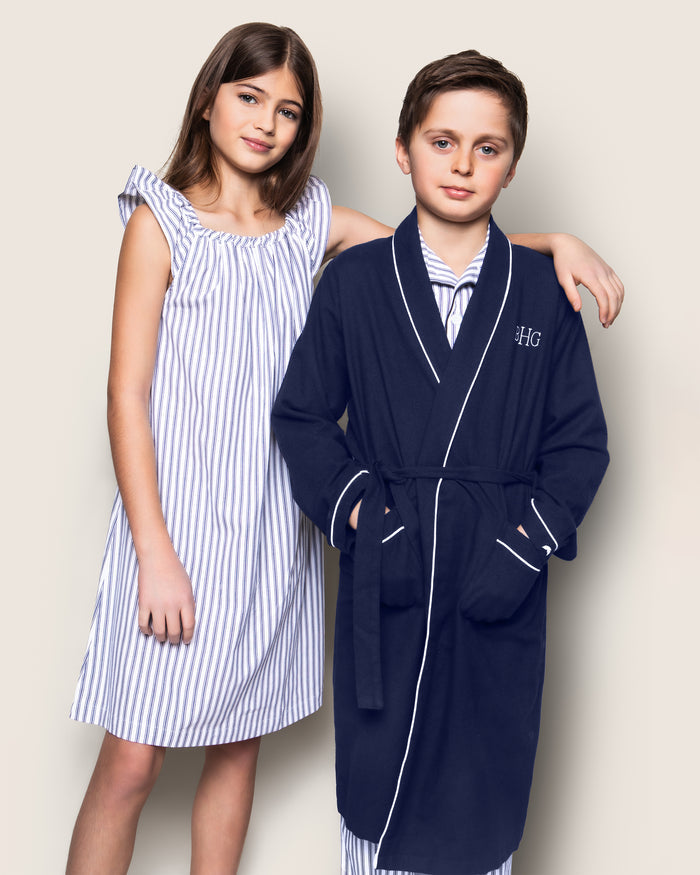 A girl in a striped dress stands next to a boy wrapped in Petite Plumes Kids Flannel Robe in Navy with White Piping, showcasing HG embroidery. Beneath, he wears flame-retardant striped pajamas. Both smile softly against a neutral background, highlighting their charming sleepwear.