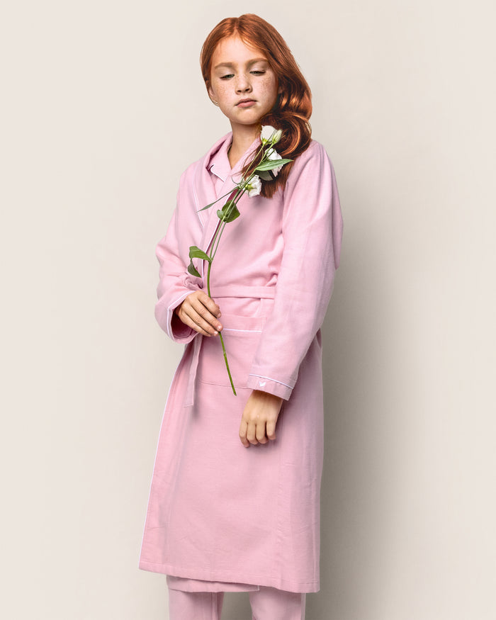 A person with long red hair stands against a plain background, wearing a Petite Plume Kids Flannel Robe in Pink. They hold a white flower gently in their hands, looking down thoughtfully.