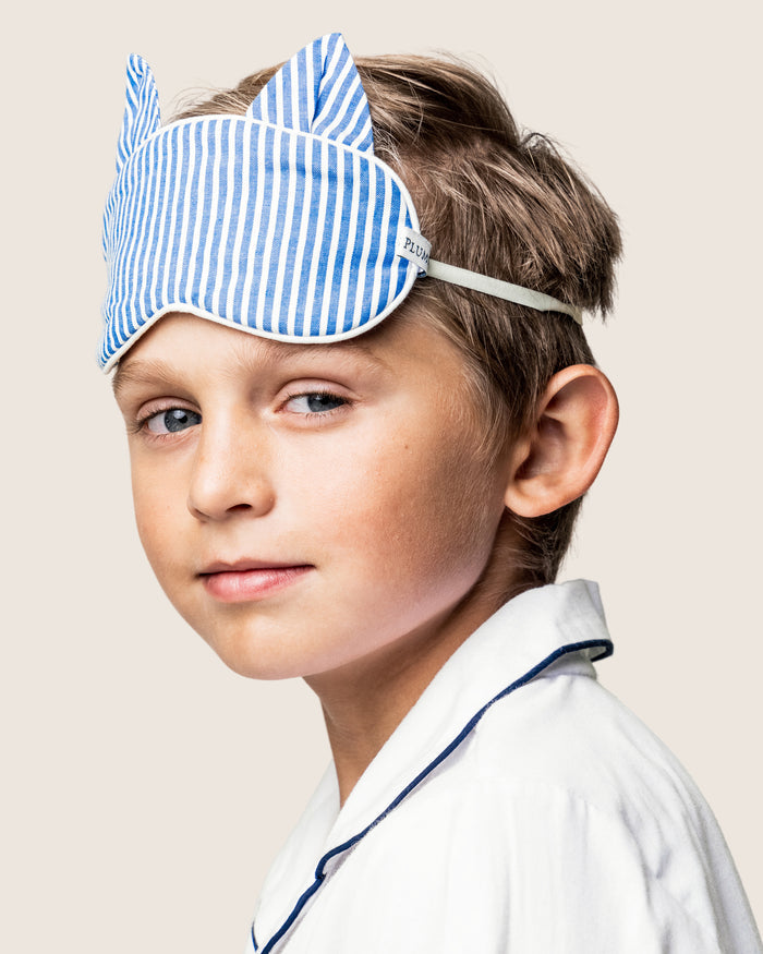 A young boy in white pajamas with dark piping stands in profile, wearing a blue and white striped Kids Kitty Sleep Mask from Petite Plume. The mask, with cat ear designs and possibly part of a monogrammed set, contrasts charmingly against the light beige backdrop.