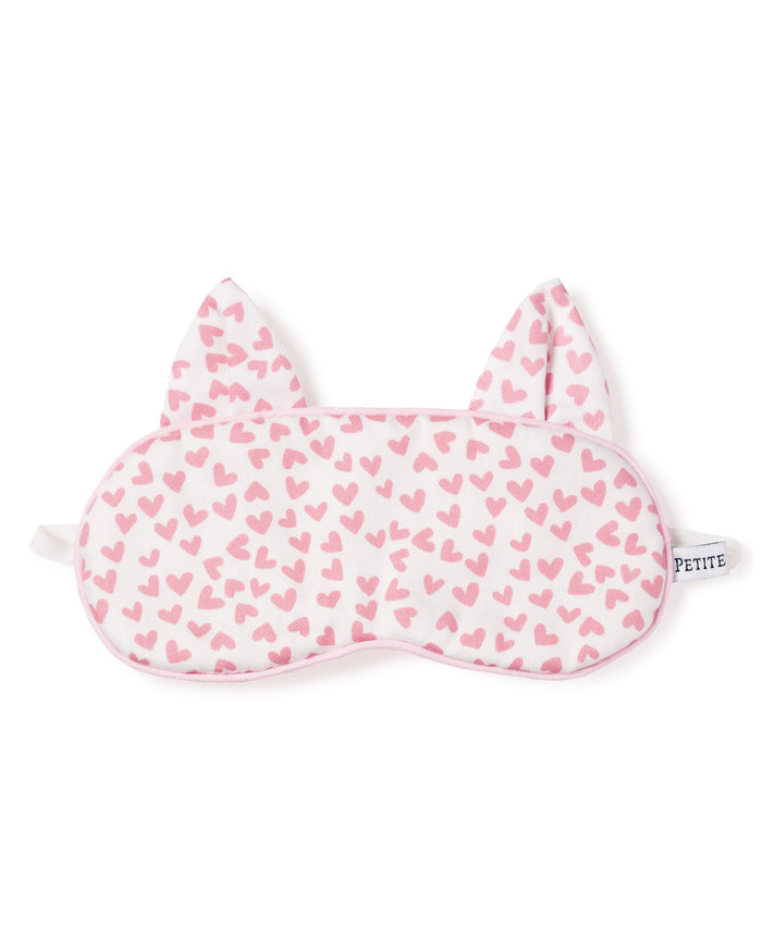 The Kids Kitty Sleep Mask in Sweethearts by Petite Plume is a charming white mask adorned with pink hearts and cute cat ears. It features side straps and a chic black label with PETITE. This luxurious accessory makes an adorable monogrammed gift.