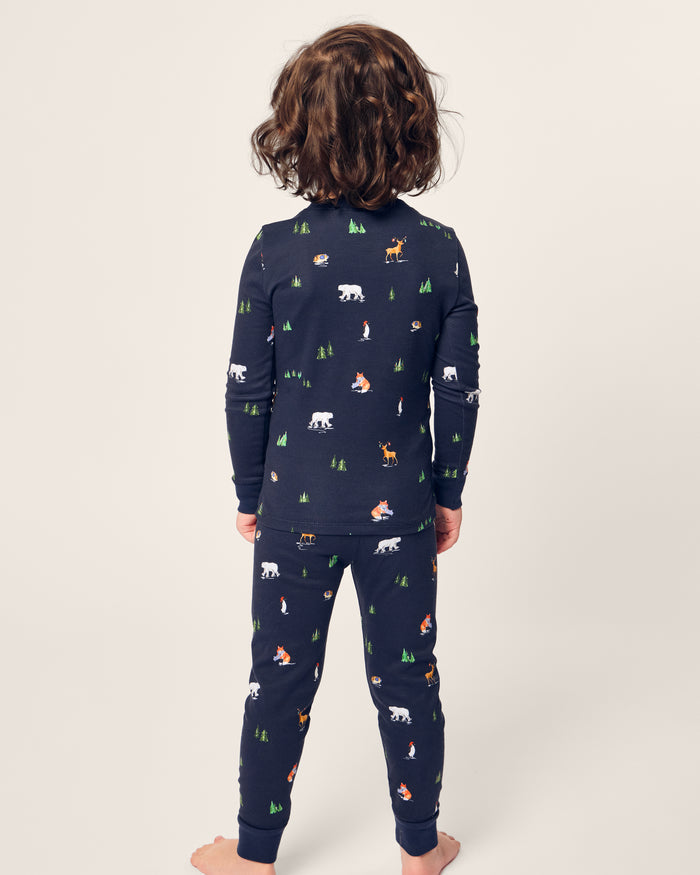 A child with curly hair stands barefoot, facing away, dressed in Petite Plumes Kids Pima Snug Fit Pajama Set in Arctic Antics. The long sleeve top and navy pajamas are made of soft Pima cotton, featuring charming bear and forest-themed motifs on a light background.