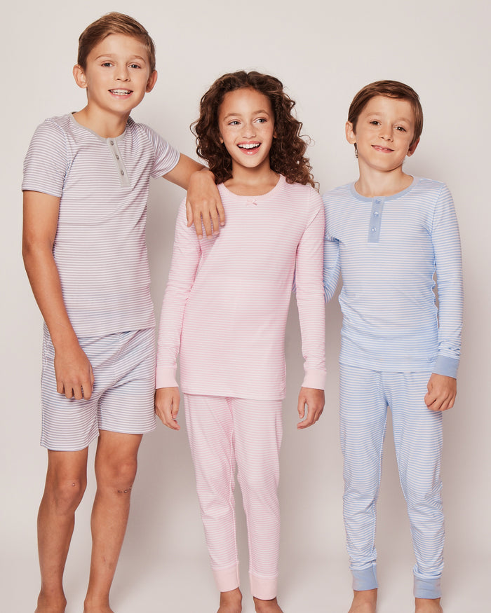 Three children smile in striped sleepwear against a plain background. The one on the left wears short sleeves and shorts, the middle in pink long-sleeves, and the right child is in Petite Plumes Kids Pima Snug Fit Pajama Set in Blue Stripes, all crafted from soft pima cotton.
