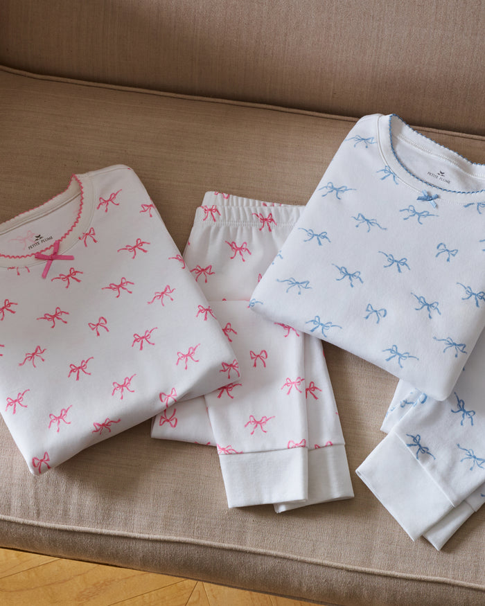 A confident young girl with long brown hair wears the Petite Plume Kids Pima Snug Fit Pajama Set in Blushing Bows, featuring a long sleeve top and pants made from soft Pima cotton with small pink patterns against a plain light-colored background.