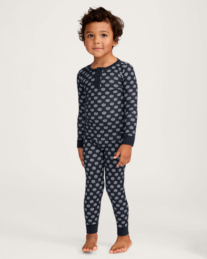 A young child with short curly hair stands barefoot against a plain light background, wearing the Kid’s Pima Snug Fit Pajama Set in Captain’s Compass—matching navy blue long-sleeve pajamas featuring a white circular pattern.