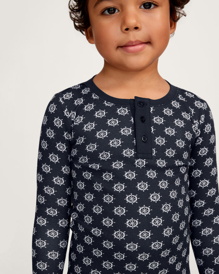 A young child wearing the Kid’s Pima Snug Fit Pajama Set in Captain’s Compass, featuring a navy blue long-sleeve top with white ship wheel patterns and a buttoned placket, stands against a plain light background.