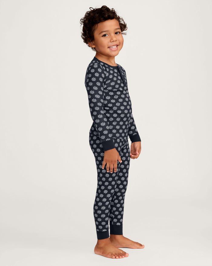 A young child with curly hair, barefoot and smiling, poses against a plain light background while wearing the Kid’s Pima Snug Fit Pajama Set in Captain’s Compass—a dark blue long-sleeve pajama set featuring a white pattern.