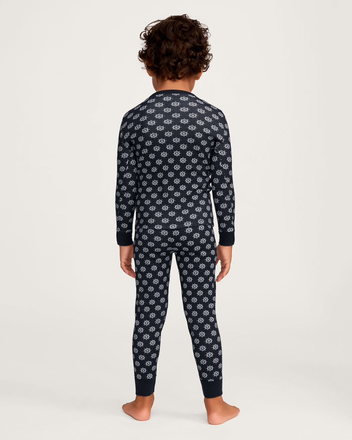 A young child with curly hair stands barefoot facing away, wearing the Kid’s Pima Snug Fit Pajama Set in Captain’s Compass, featuring matching long-sleeve top and pants with a small white geometric pattern on a dark background.