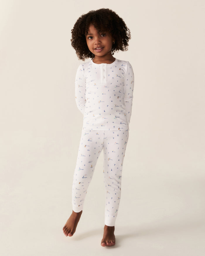 A young child with curly hair stands barefoot and smiling, hands behind their back, wearing the Kid’s Pima Snug Fit Pajama Set in Letters and Lullabies—white long-sleeve pajamas featuring small colorful prints—posing against a plain light background.