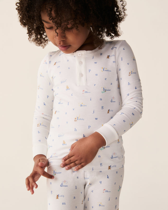 A child with curly hair wears the Kid’s Pima Snug Fit Pajama Set in Letters and Lullabies, featuring white fabric decorated with small, colorful letters and words. The child looks down while gently pulling at the hem of their shirt.